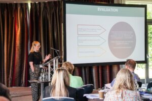 Jess Smith, Research and Evaluation Manager at Framework talking at the Under One Roof Conference 2026. She is pointing at her presentation which is projected on a big screen in front of an audience.