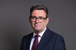 A portrait of Andy Burnham. He is wearing a navy suit and a deep red tie with a white shirt.