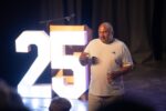 Adam standing in front of the audience at YSA 2026 talking. He is stood in front of a sign that reads 25 and it isglowing.