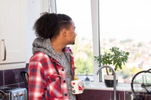 A man holding a white mug and looking out the window. He looks serious and is wearing a grey hoodie with a red checkered jacket.